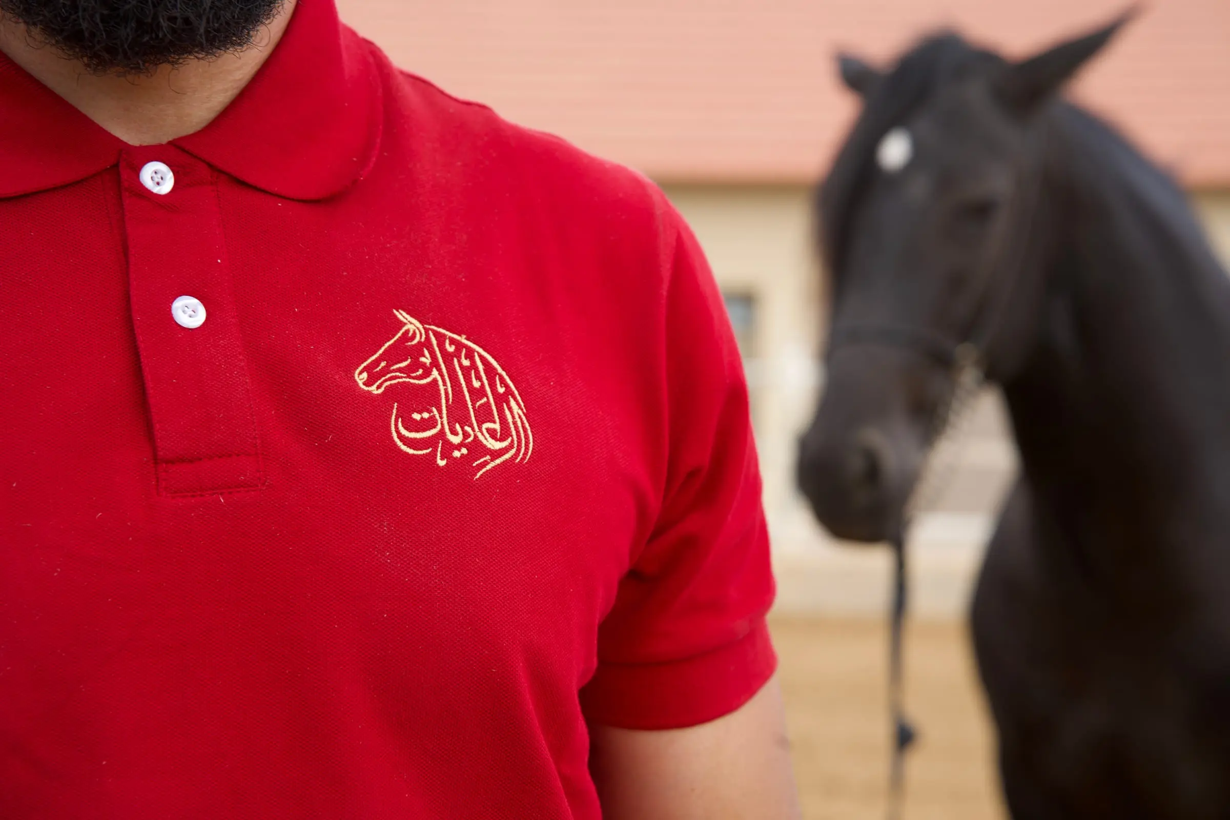 Polo rouge brodé du logo El Adiyat, porté par un palefrenier, cheval noir en arrière-plan